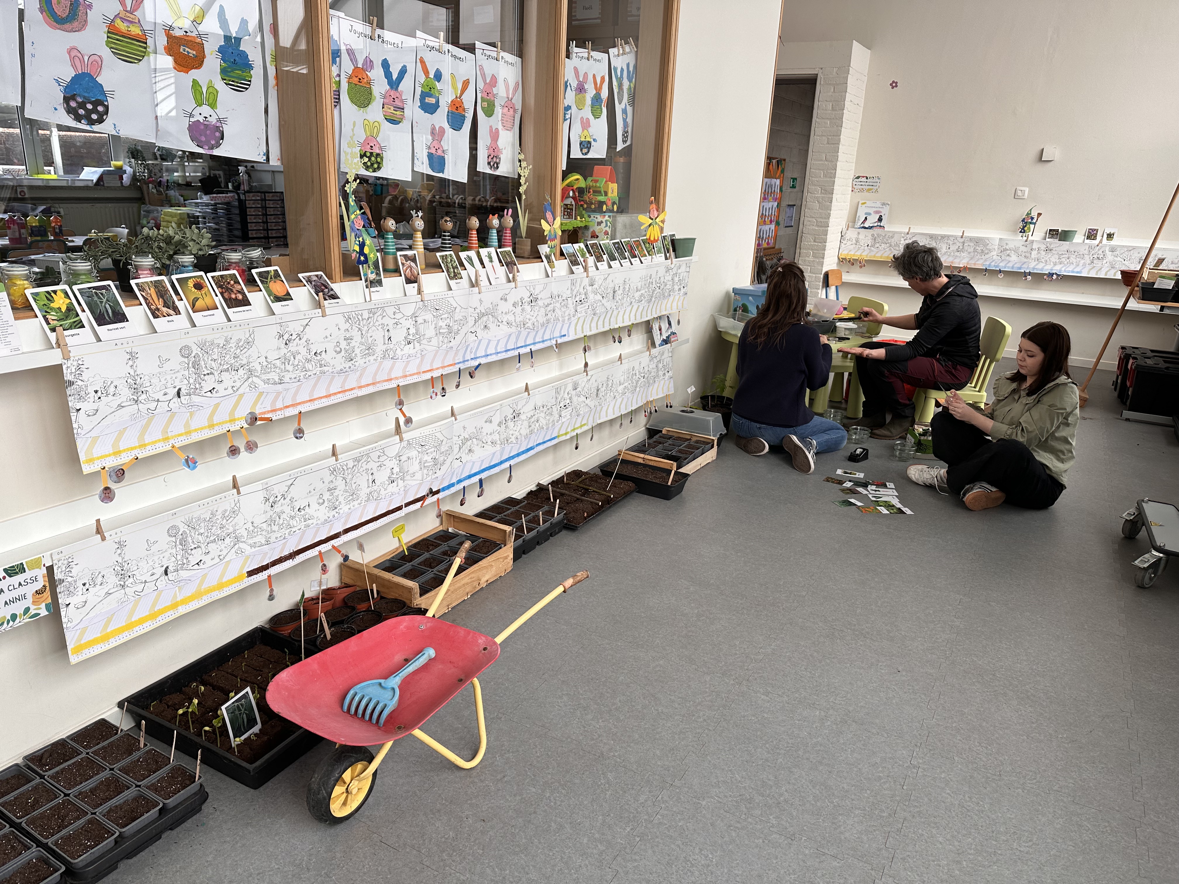 Le Calendrier Panoramique affiché au mur en 1ère maternelle du CSM, avec les bacs à semis au sol et les institutrices préparant les plantations