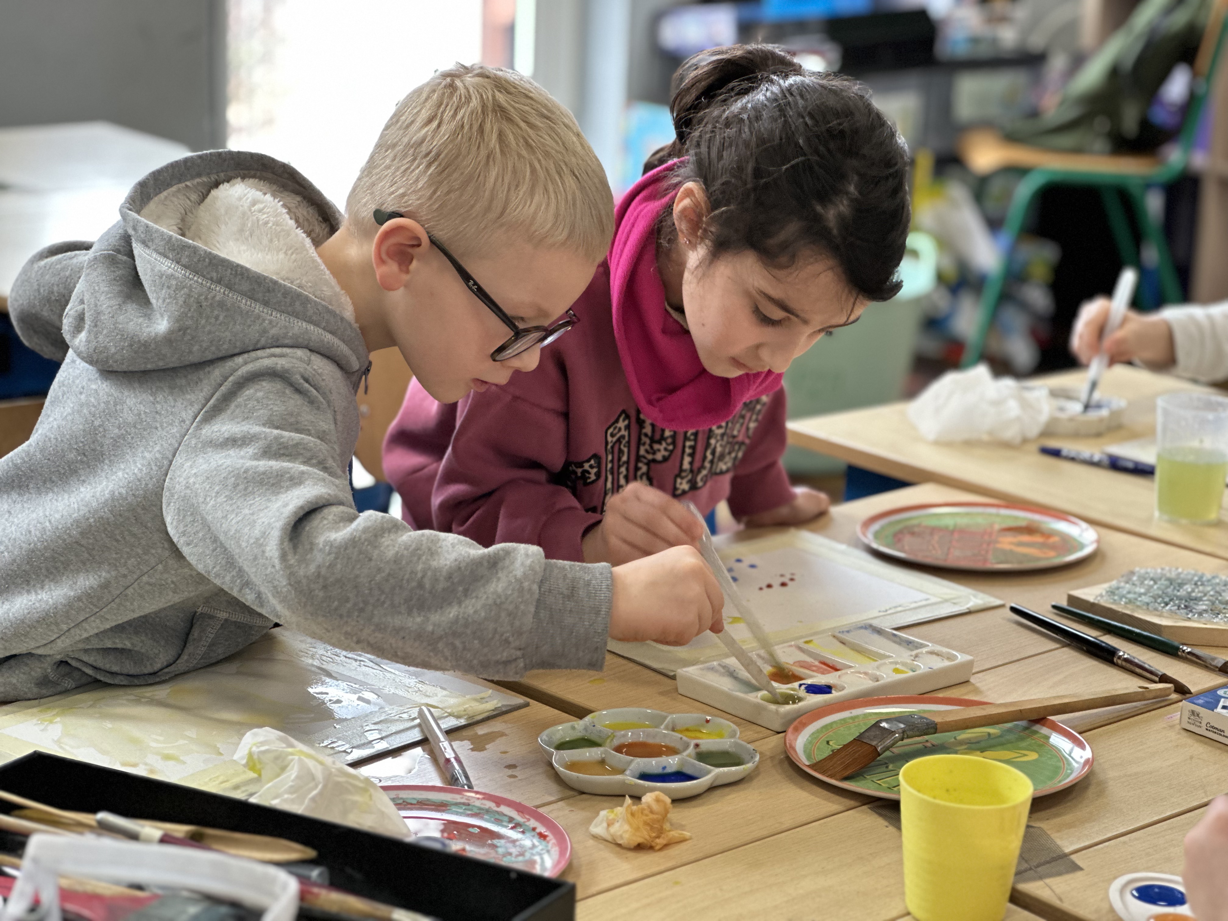 Une fille et un garçon penchés sur leur palette d'aquarelle, pipettes en main, concentrés