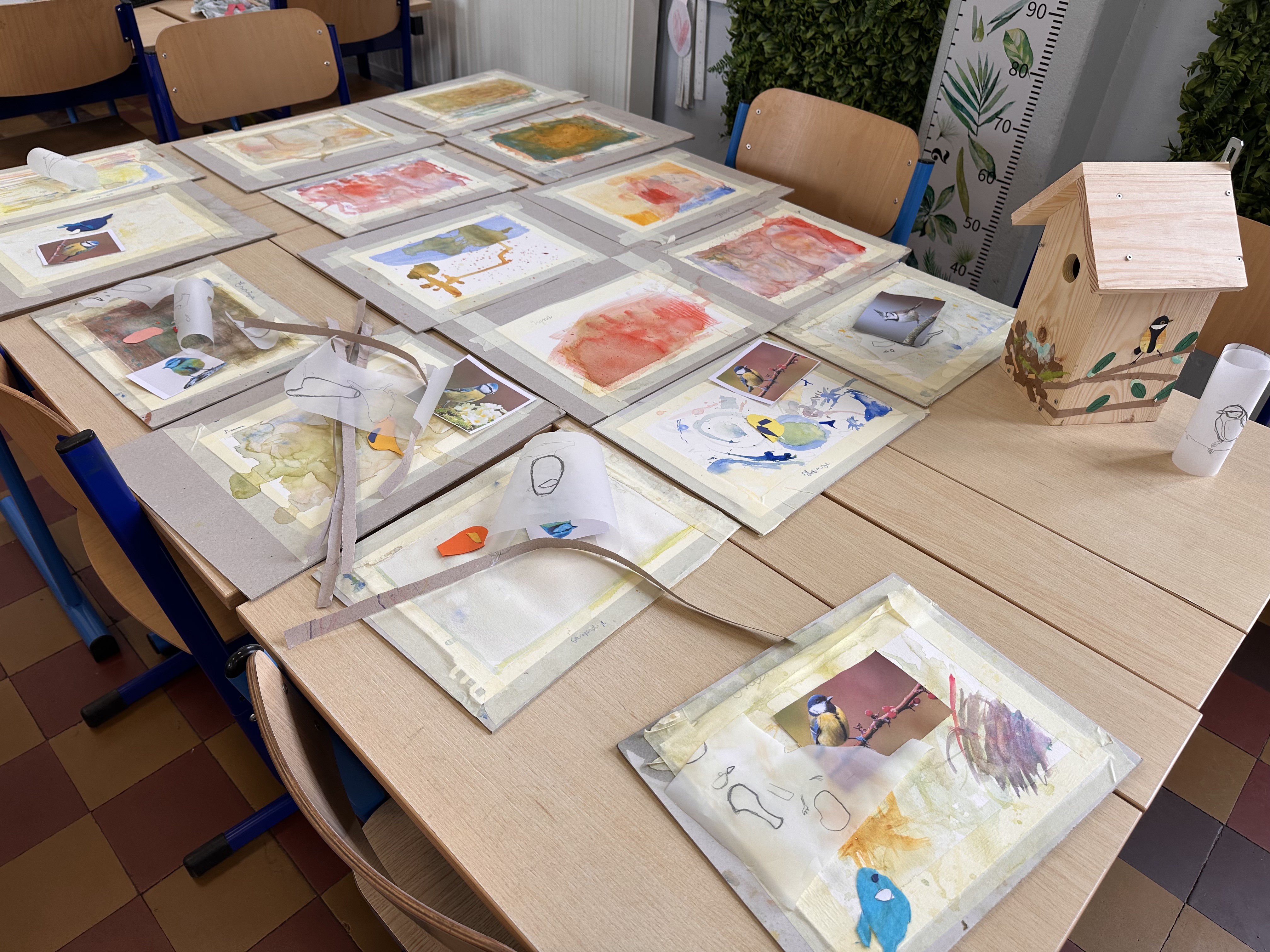 Petites œuvres aquarelle des enfants de 2ème primaire posées sur un bureau, prêtes pour le collage